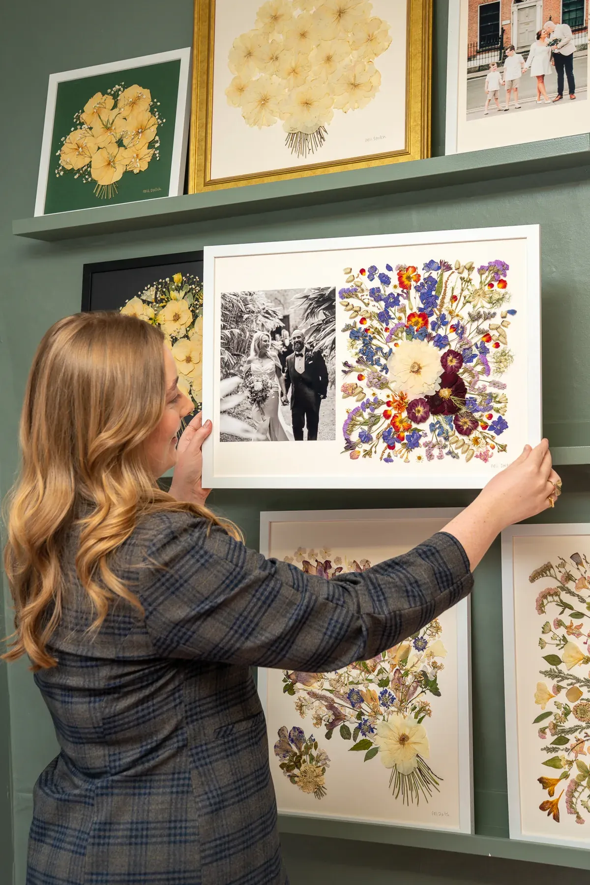 Nell hanging a framed pressed bouquet on the wall beside a wedding photograph