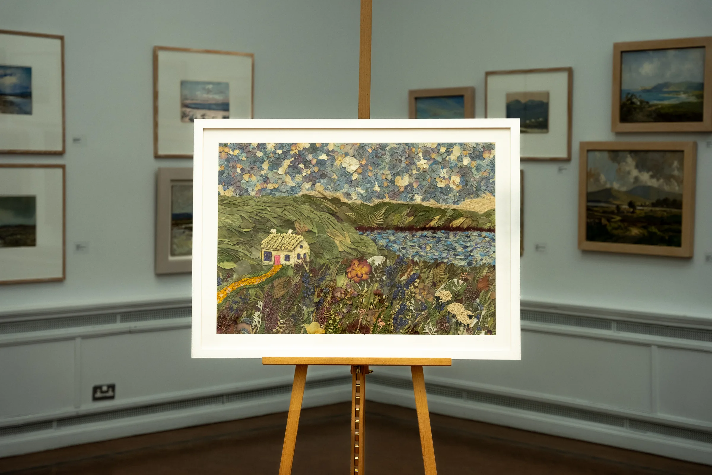 A pressed flower cottage landscape on an easel in a gallery