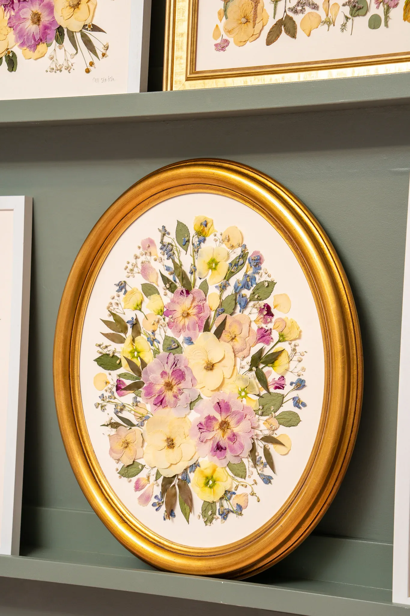 A pressed bouquet in a gold oval frame on the studio shelf