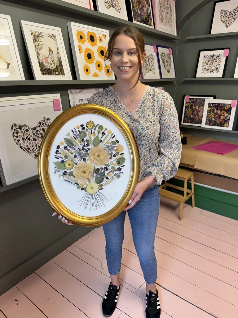 A smiling customer holding her framed pressed flower piece in a gold oval frame