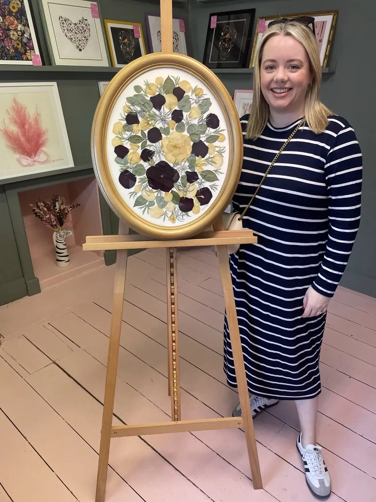 A customer smiling beside her pressed flowers in a gold oval frame on an easel