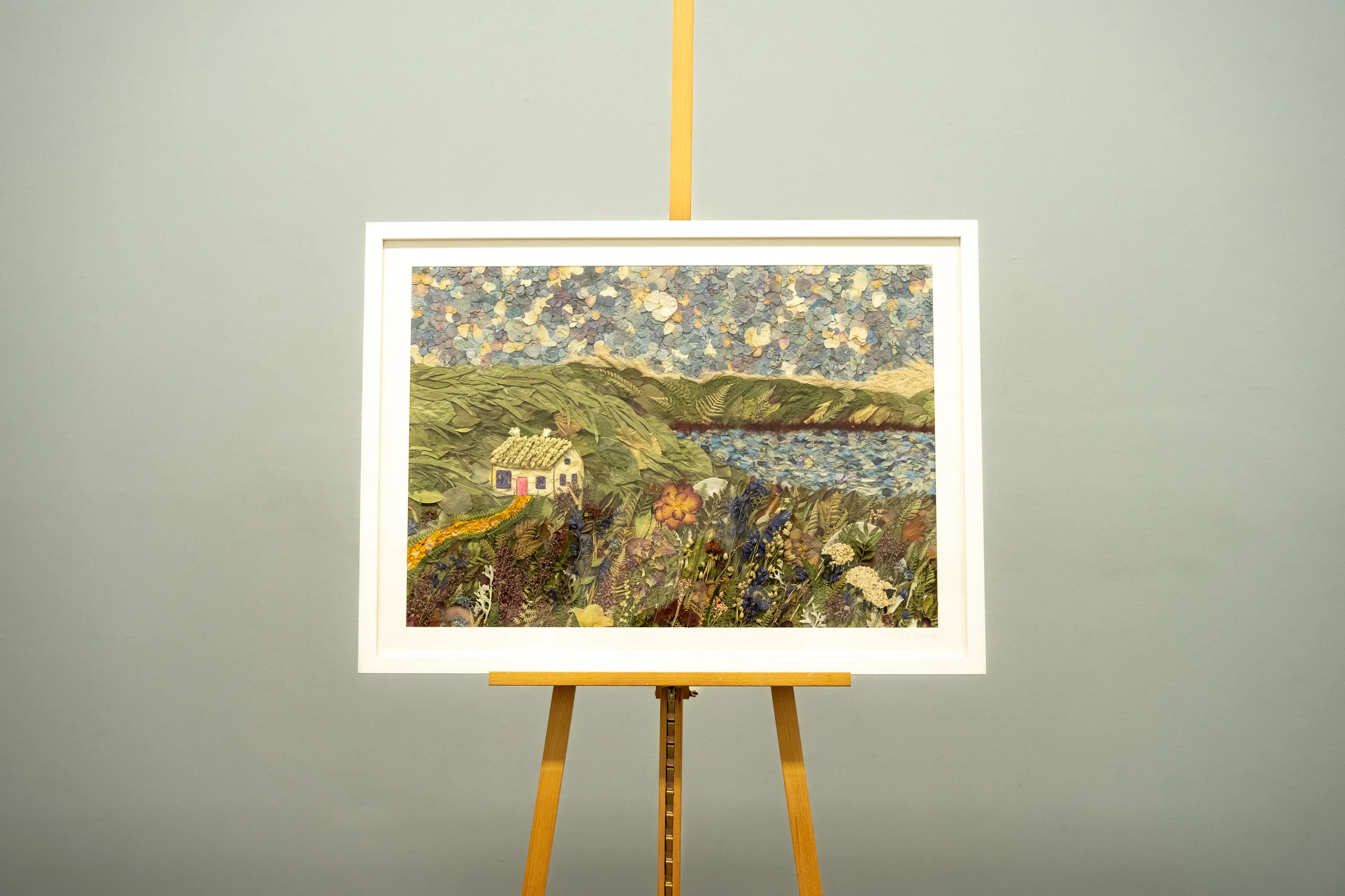 A bespoke pressed flower landscape of a cottage by a lake, in a white frame on an easel